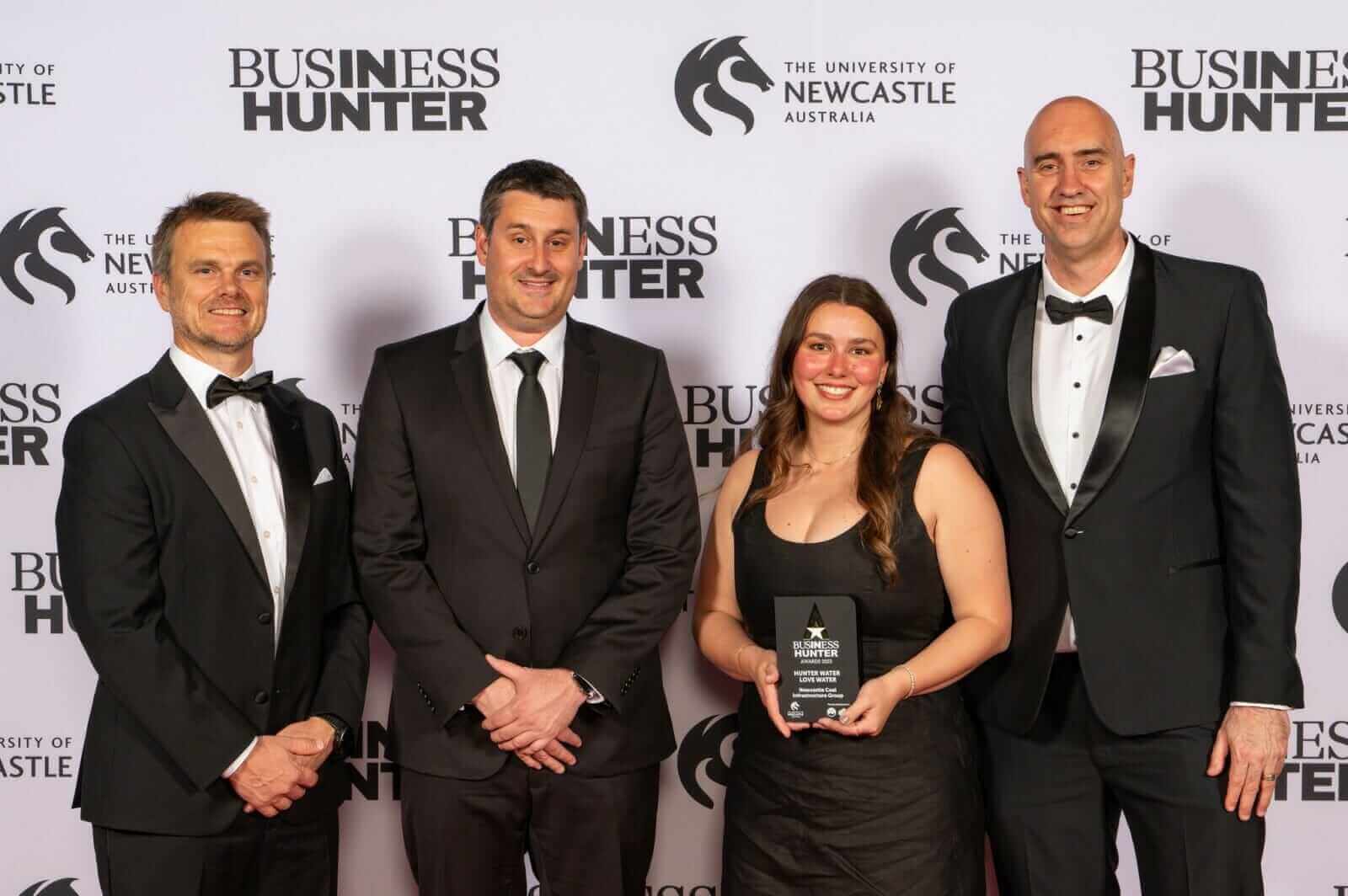 Three men in black suits and a lady in a black dress stand in front of a media wall with an award.